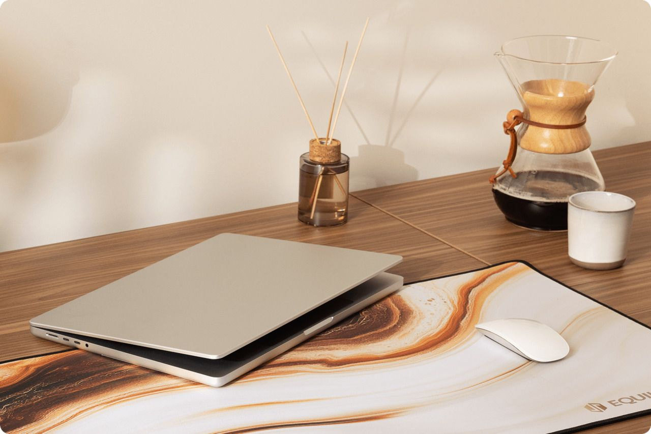 A wooden desk featuring a laptop, mouse, coffee cup, and a vase of flowers, creating a cozy workspace atmosphere.