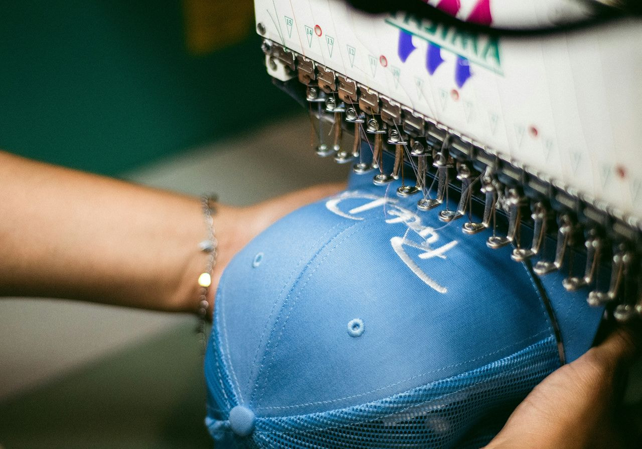 A blue cap is being embroidered by a machine, with white thread forming intricate patterns.