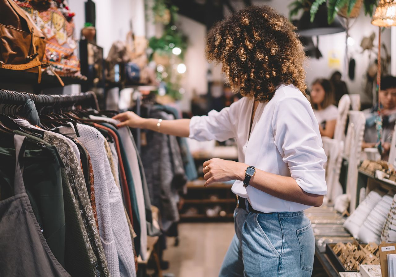 A woman in a white shirt and jeans browses a rack of secondhand clothing in a vintage-style clothing store.