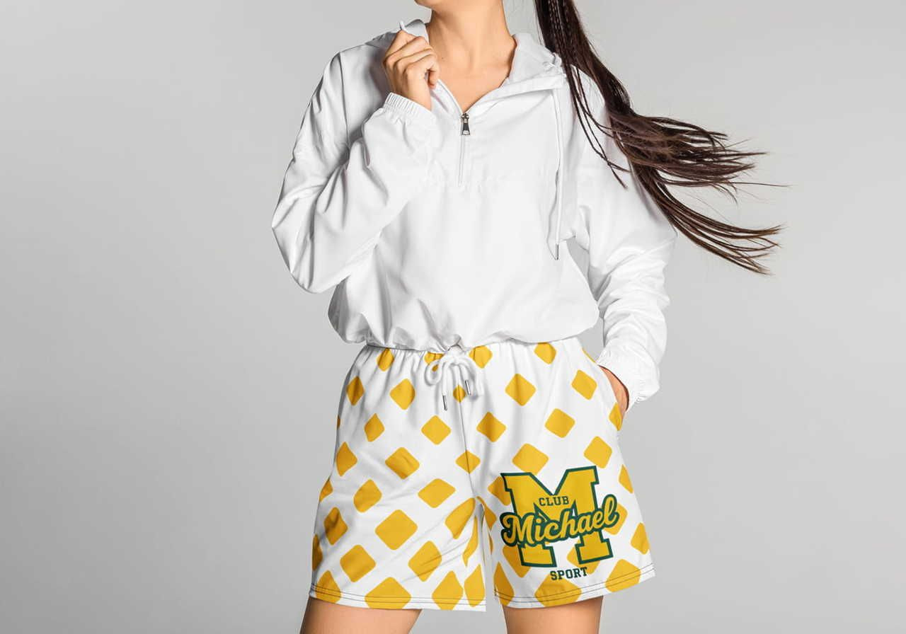 A woman in a white jacket poses with her hand near her face, wearing white shorts with yellow diamond patterns and a green "Club Michael Sport" logo.