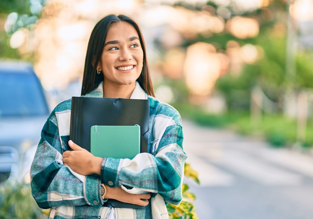 Smiling young woman standing outdoors, holding books and a folder. 