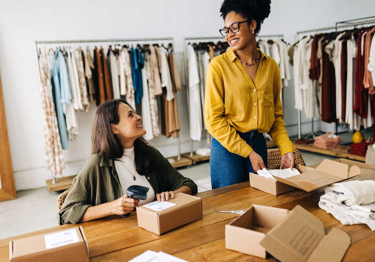 Two women happily pack boxes in a clothing store, surrounded by racks of colorful outfits. One holds a barcode scanner.