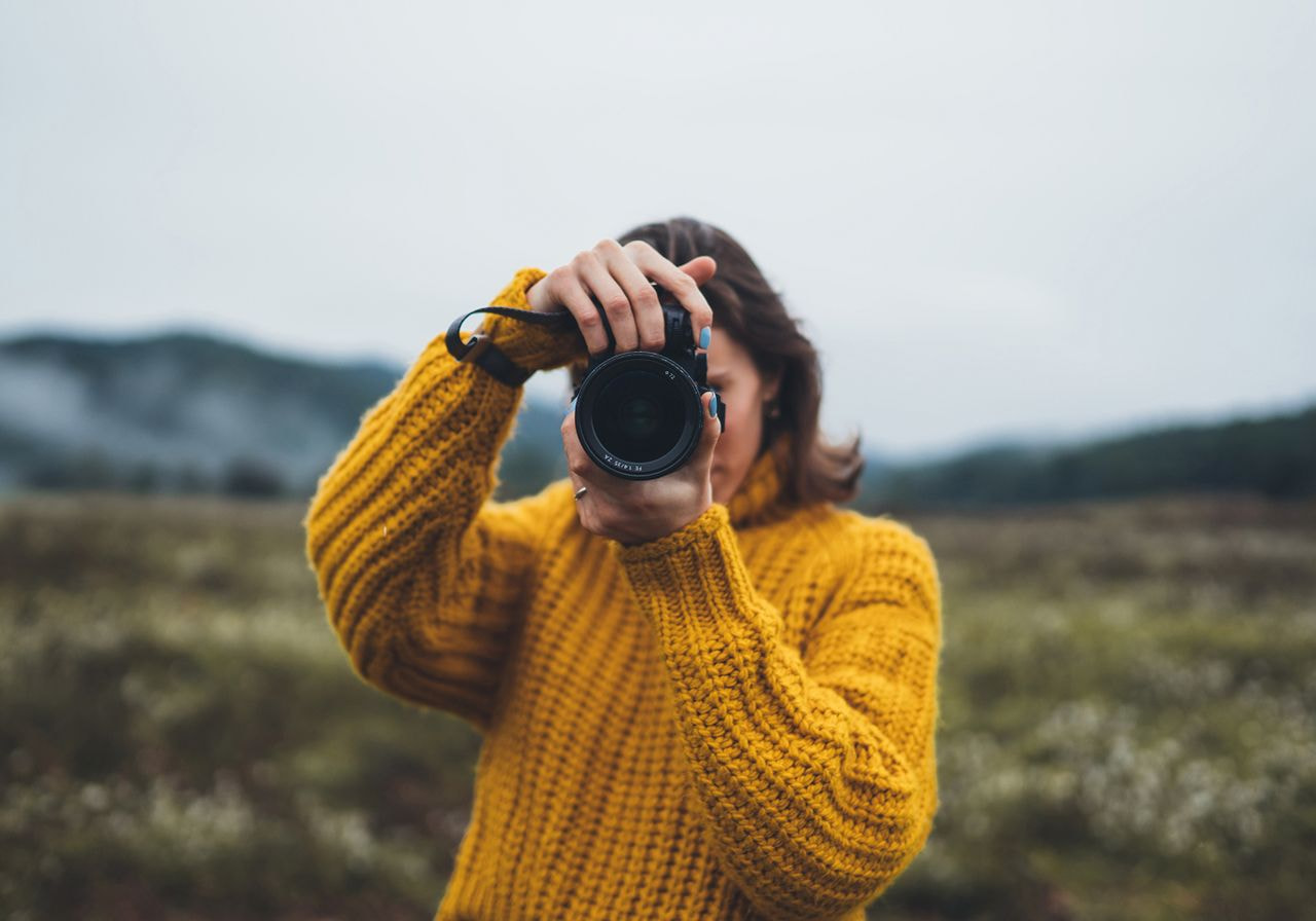 A person in a yellow sweater takes a photo with a camera, standing in a misty field with distant mountains. 
