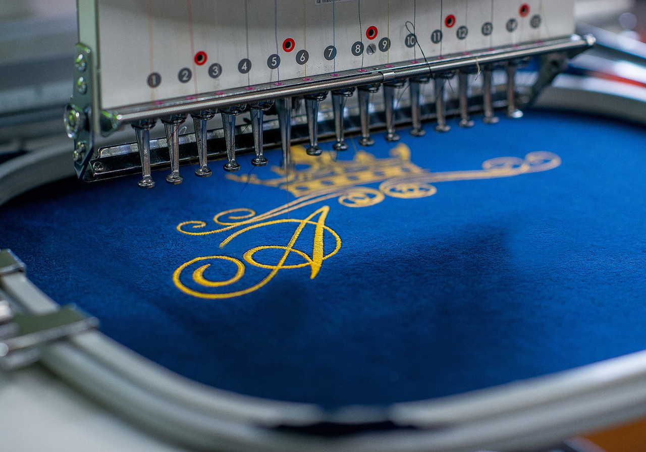 Embroidery machine stitching a golden crown and ornate letter "A" on blue fabric.