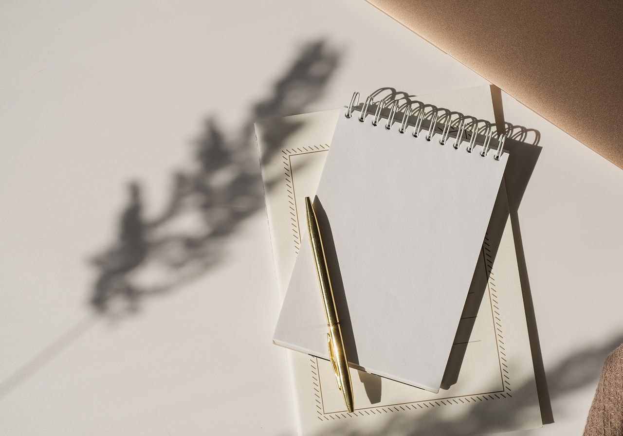 A blank notepad and pen placed on a white table next to an open notebook.