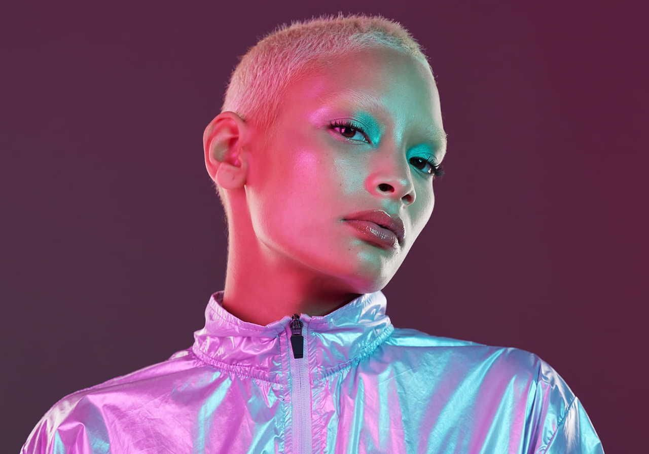 A woman with short platinum hair poses in a shiny silver jacket against a gradient purple background.