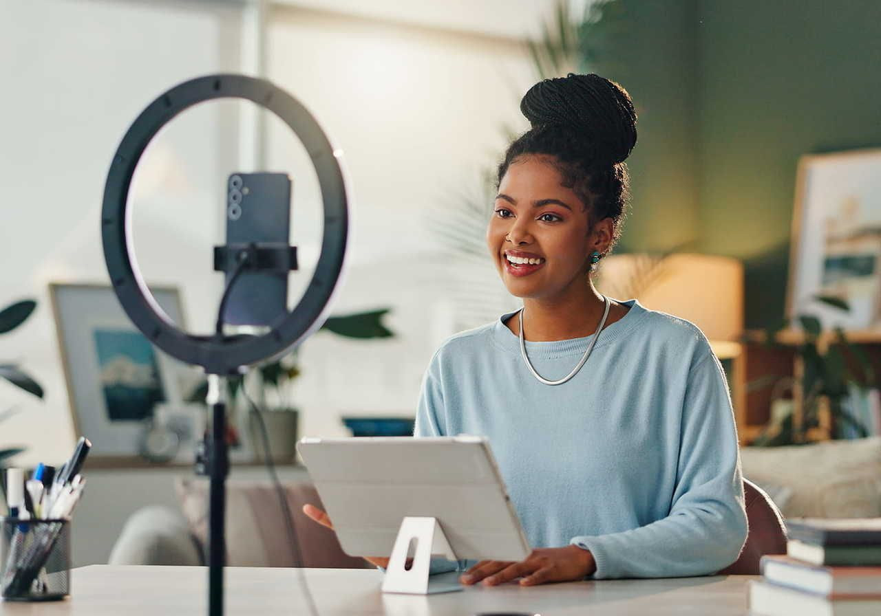 A young woman sits at a desk, smiling while using a tablet, with a ring light and a smartphone on a stand in front of her.