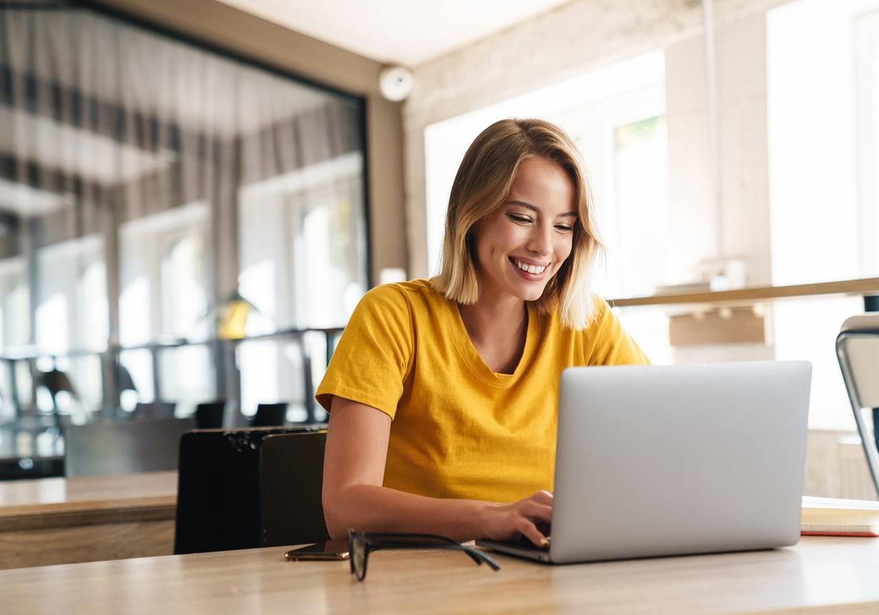 A woman in a yellow shirt sits at a table, working on a laptop.