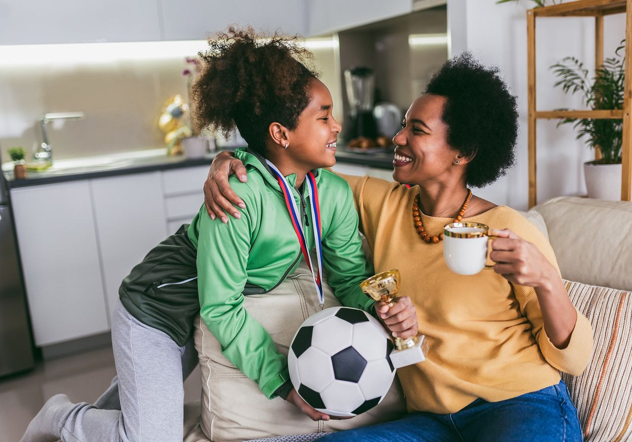 Mother and daughter sit on a couch, smiling while holding a soccer ball and a trophy cup between them.