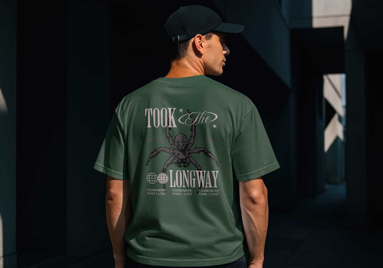 A man in a dark cap stands in an urban setting, wearing a green t-shirt with a spider graphic and the words "Took the long way”.