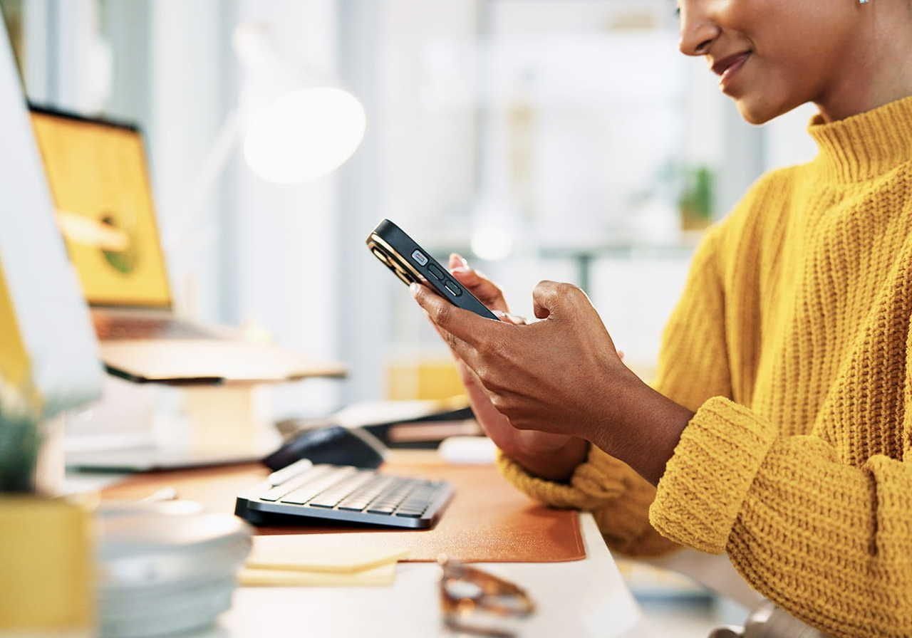 A woman in a yellow sweater is sitting at a desk, holding a phone and looking at it, with a computer and other office items in the background.
