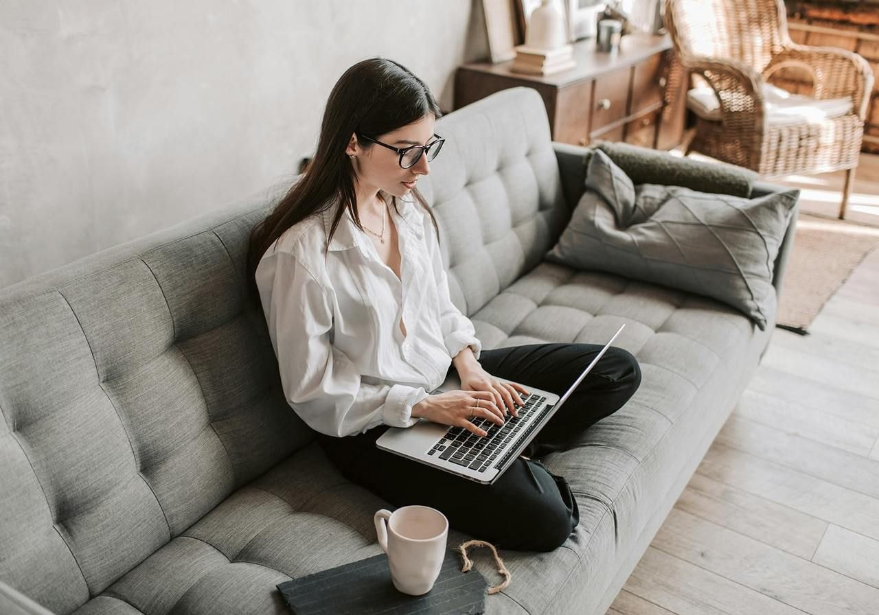 A young woman in glasses is siting on a couch and using a laptop.