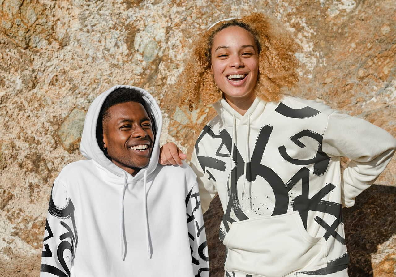 Two people smile and pose against a textured rock background. They wear white hoodies with bold, abstract black designs.