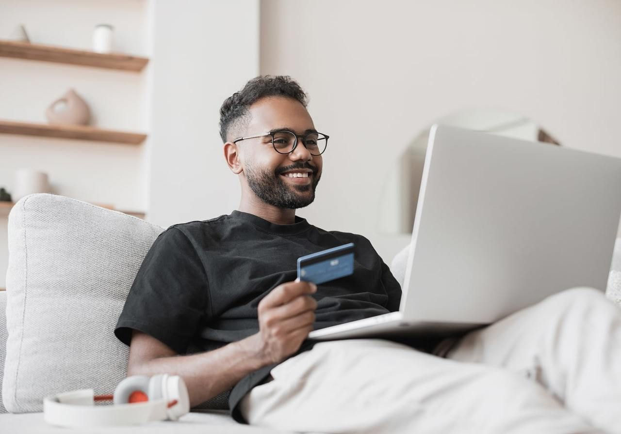 A smiling man with glasses holding a credit card to make a Twitch donation, using a laptop on a cozy couch.