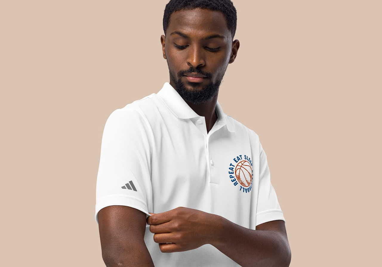 A man in a white polo shirt with a basketball emblem adjusts his sleeve against a beige background.