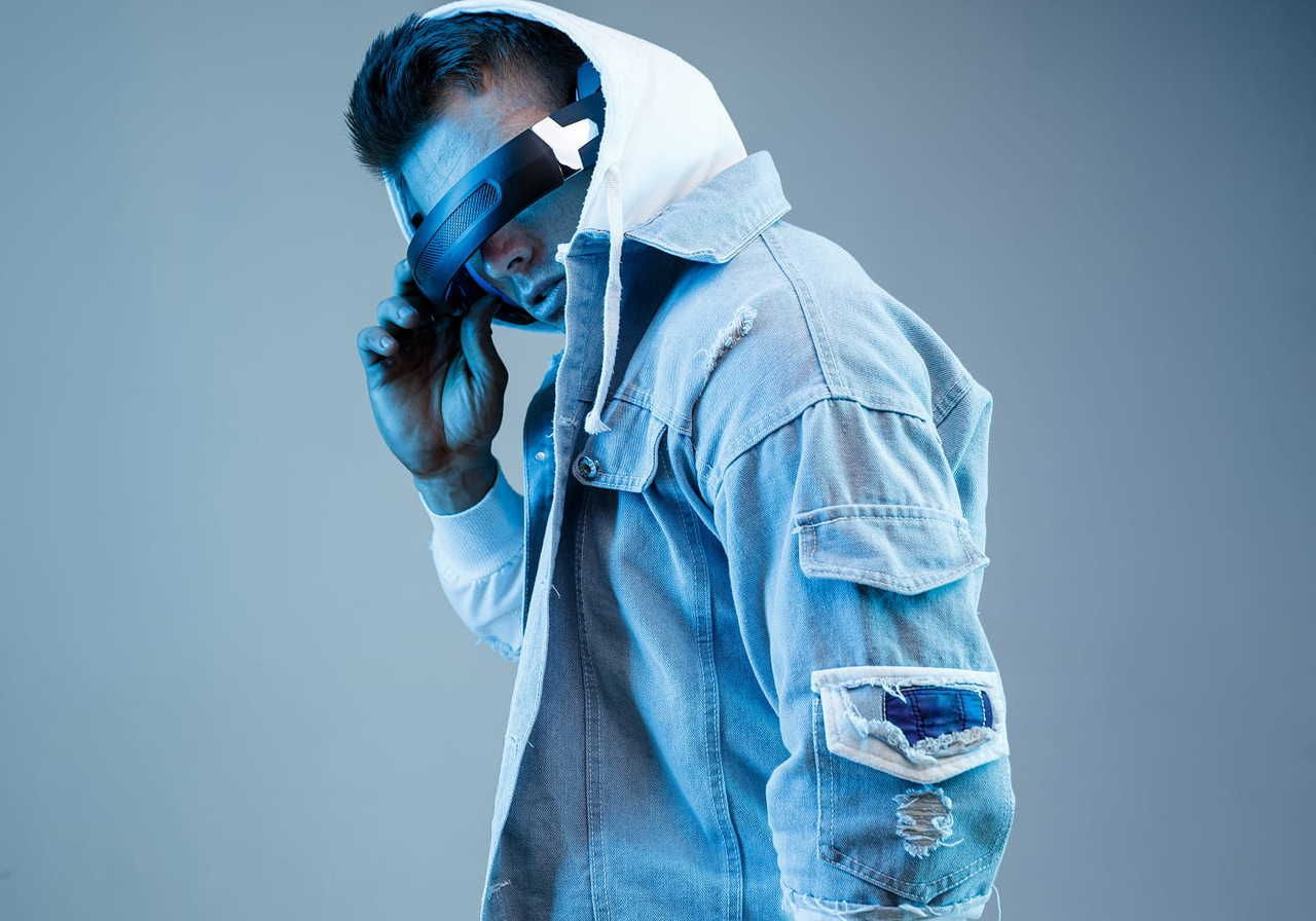 A man wearing futuristic glasses is in a white hoodie and jeans. He is looking directly into the camera.