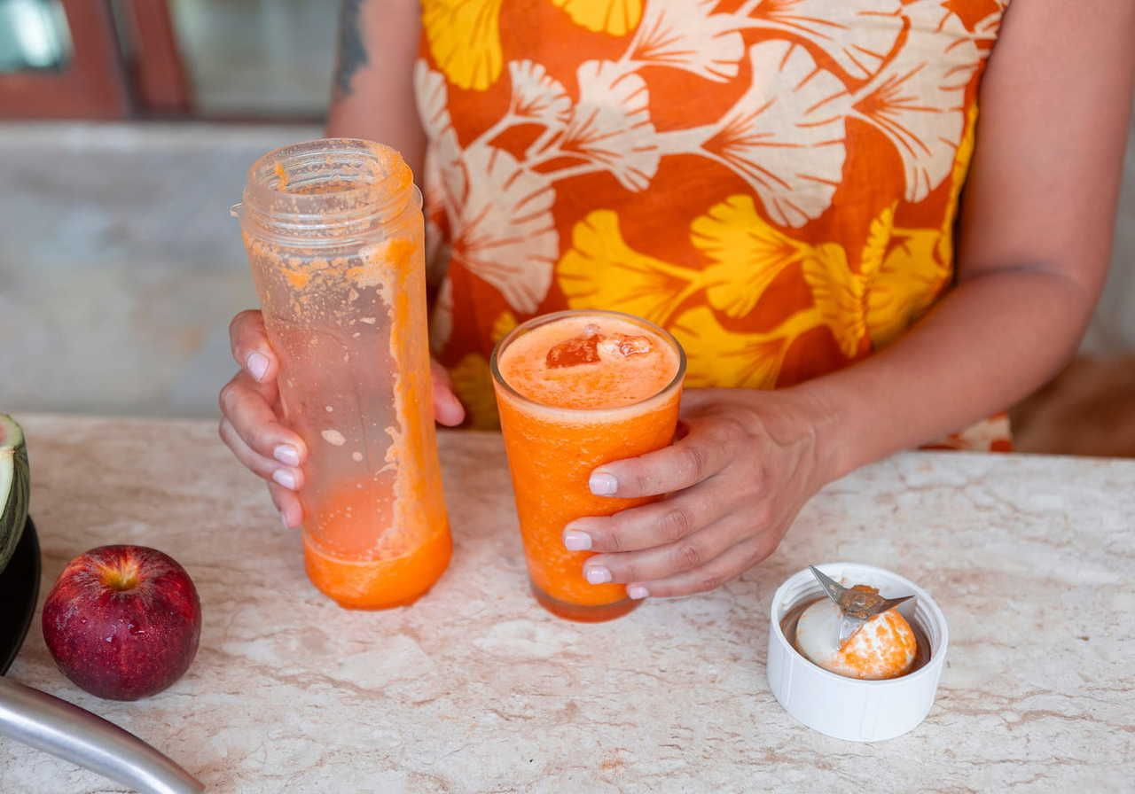 A woman holding a blender and a glass of orange smoothie on a countertop, with an apple and a small bowl on the side.