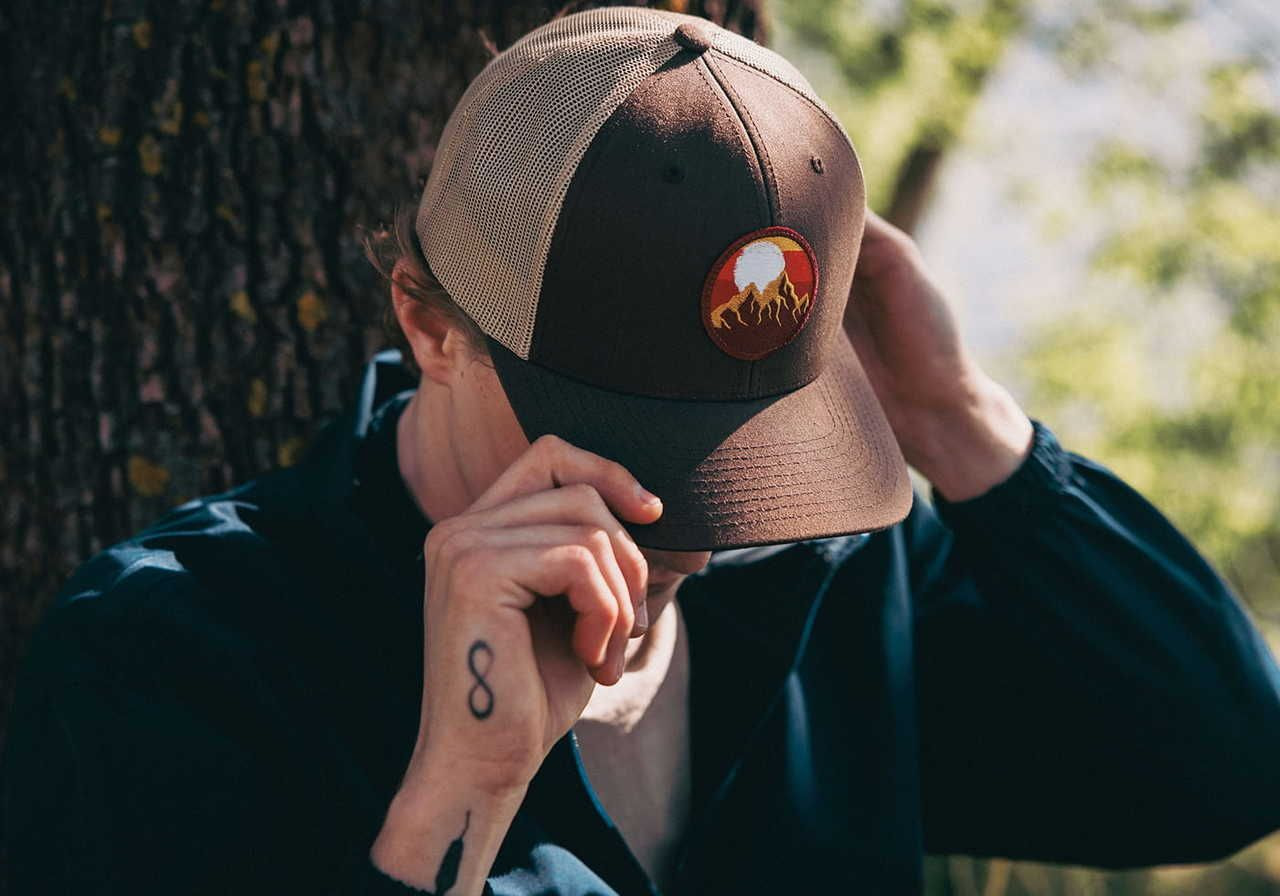 A man wearing a brown hat with a logo of a sunset and mountains, adjusting the hat while standing under a tree.