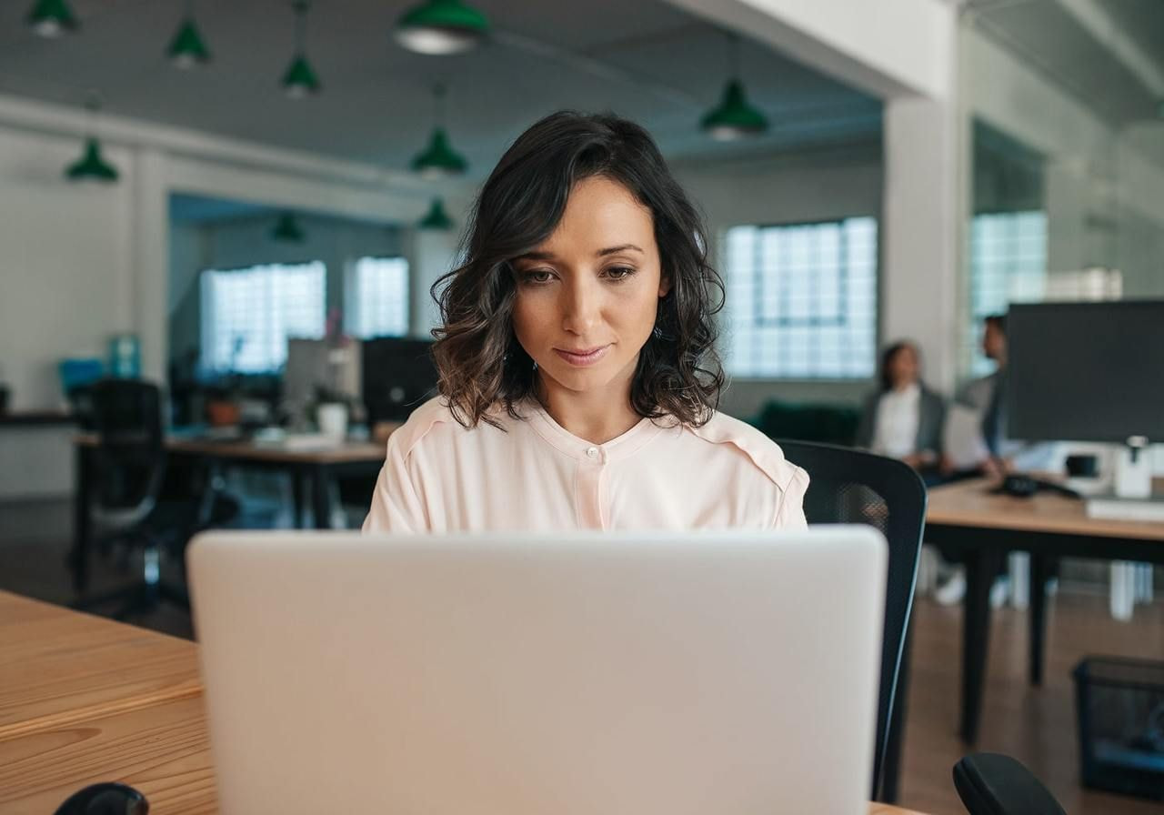 A woman focused on a laptop in an open-plan office. Two colleagues are in the blurred background.