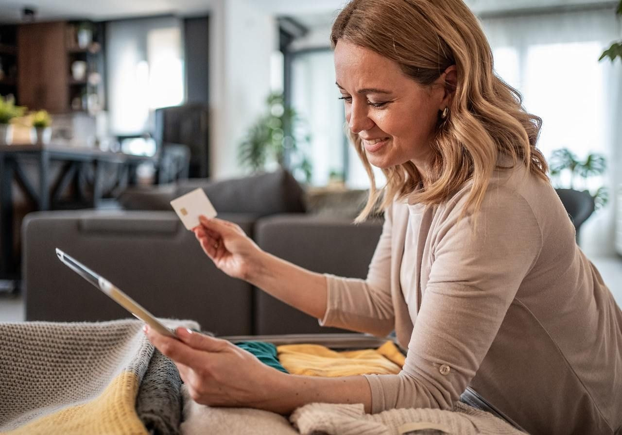 A smiling woman shops online from home, holding a credit card and a tablet.