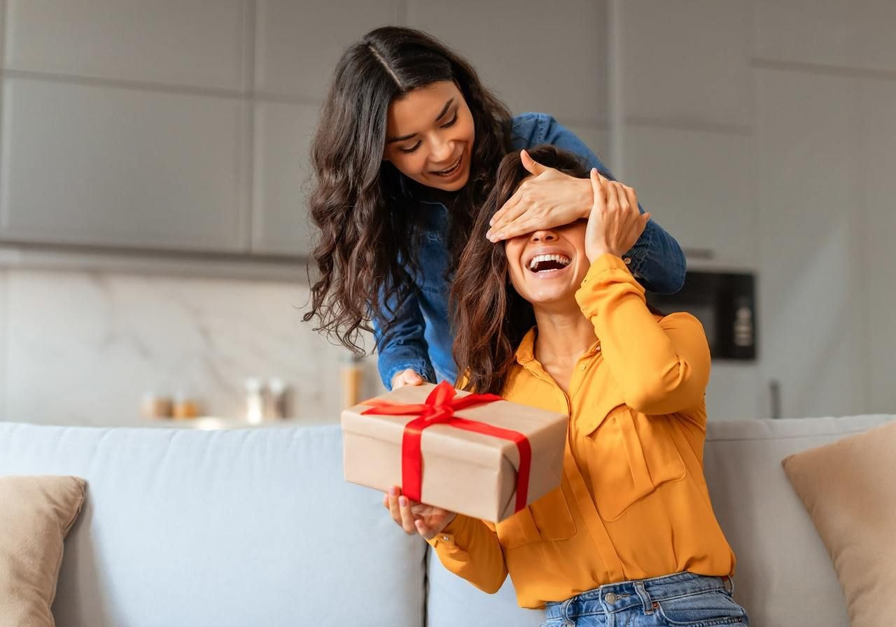 A woman in a yellow blouse smiles while holding a gift box with a red ribbon. Another woman playfully covers her eyes.