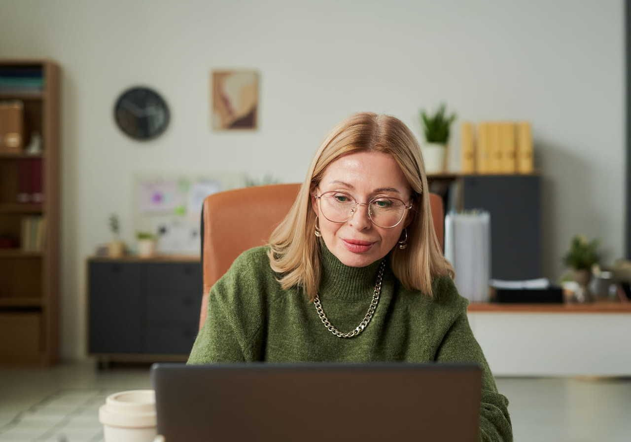 A woman with glasses and a green sweater sits at a desk, focused on a laptop. Shelves with books and a plant in the background.