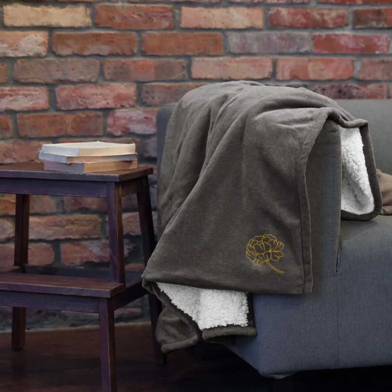 A cozy, dark gray blanket with a white, fluffy interior and yellow floral embroidery drapes over a gray sofa, beside a small wooden table.