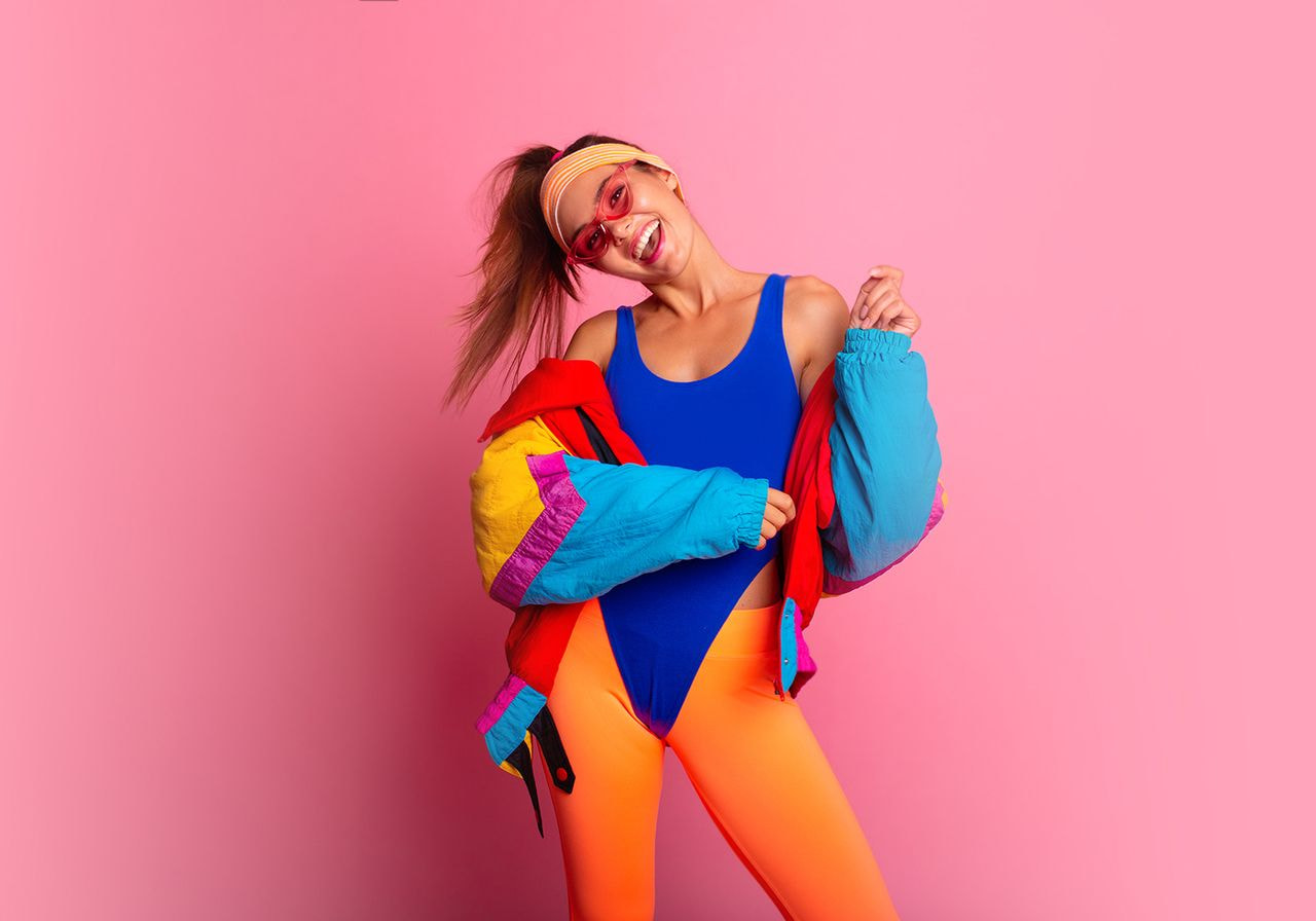 A woman in a blue leotard and orange leggings poses cheerfully against a pink background.