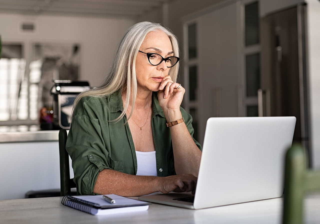A woman with long, gray hair and glasses sits at a table with a laptop, looking thoughtfully at the screen. A notebook is placed beside her.