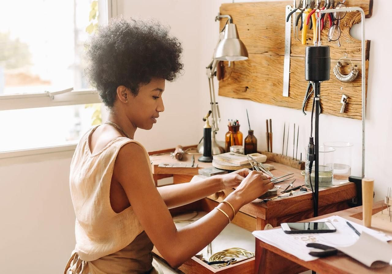 A focused individual sits at a craftsman's workbench, surrounded by tools and materials, carefully assembling jewelry.