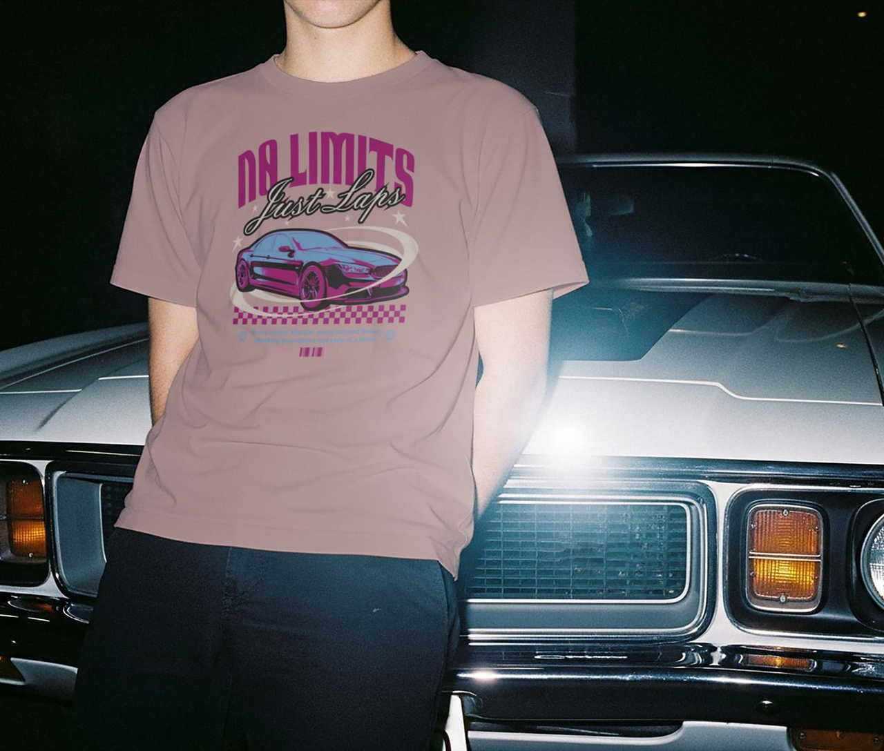 A man leans against a vintage car at night, wearing a dark-pink t-shirt with a racing-car graphic and the words "No Limits, Just Laps".