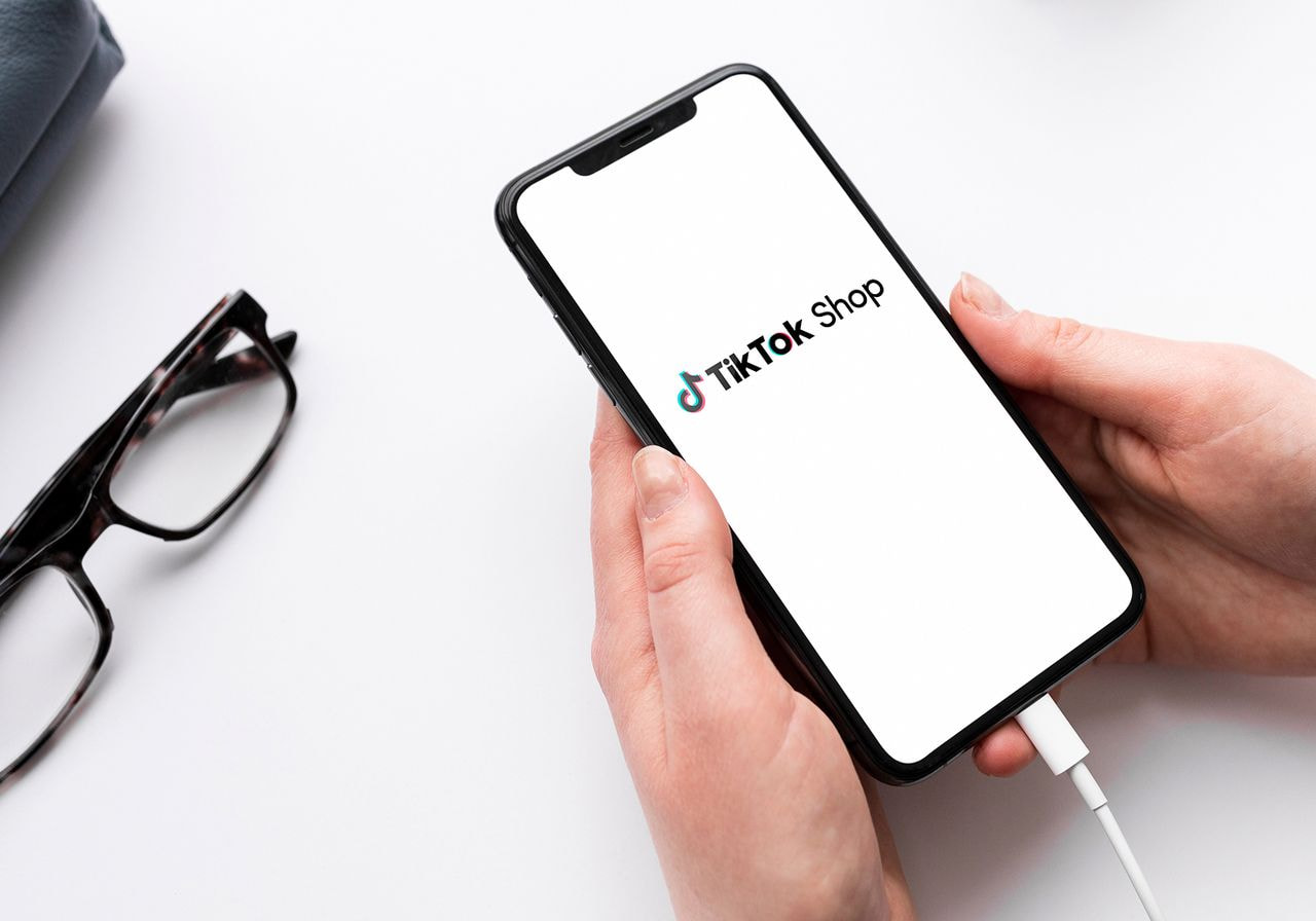 A person is holding a smartphone displaying the "TikTok Shop" logo on a white screen.