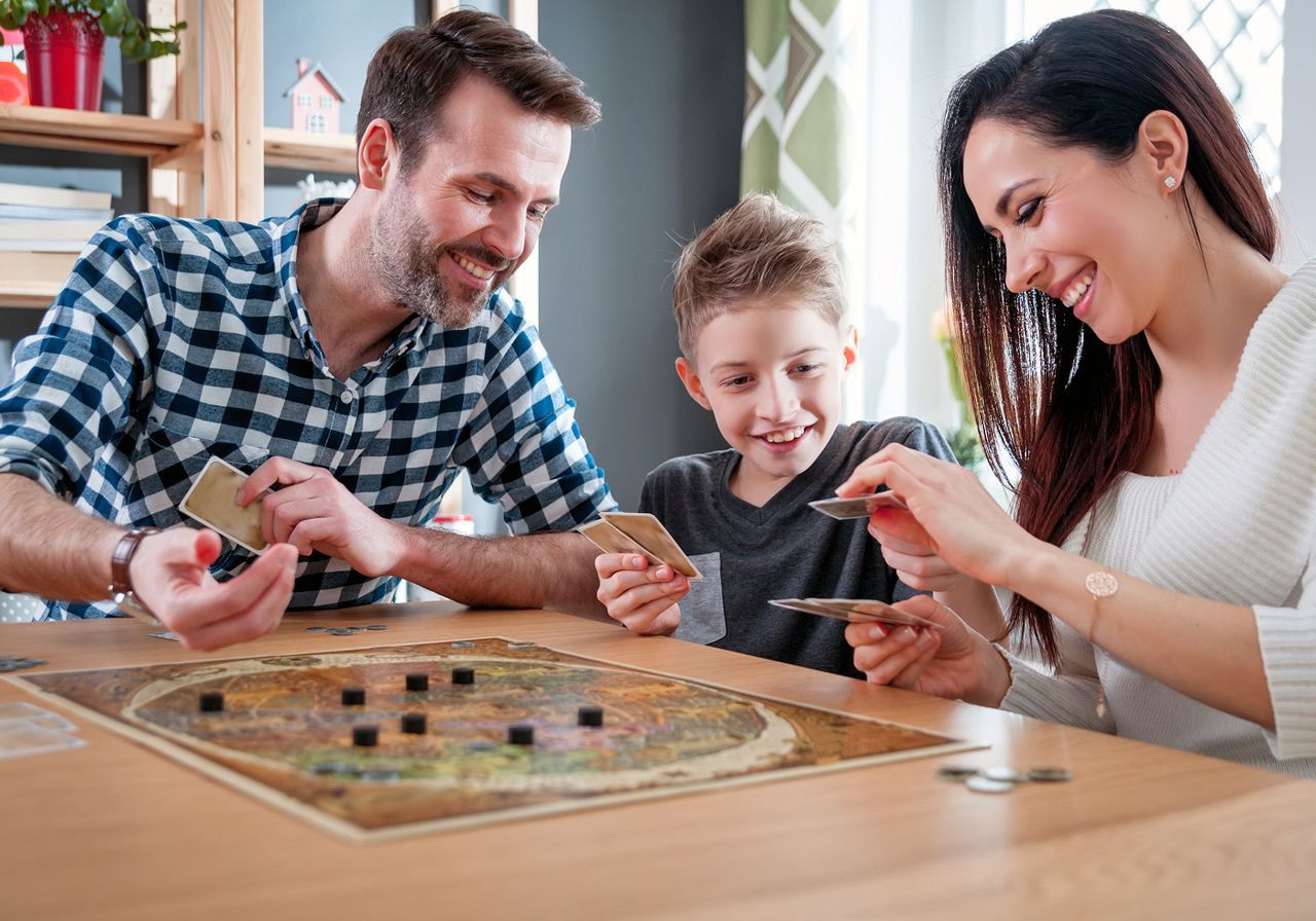 A smiling family of three plays a board game with cards at a wooden table in a bright living room.