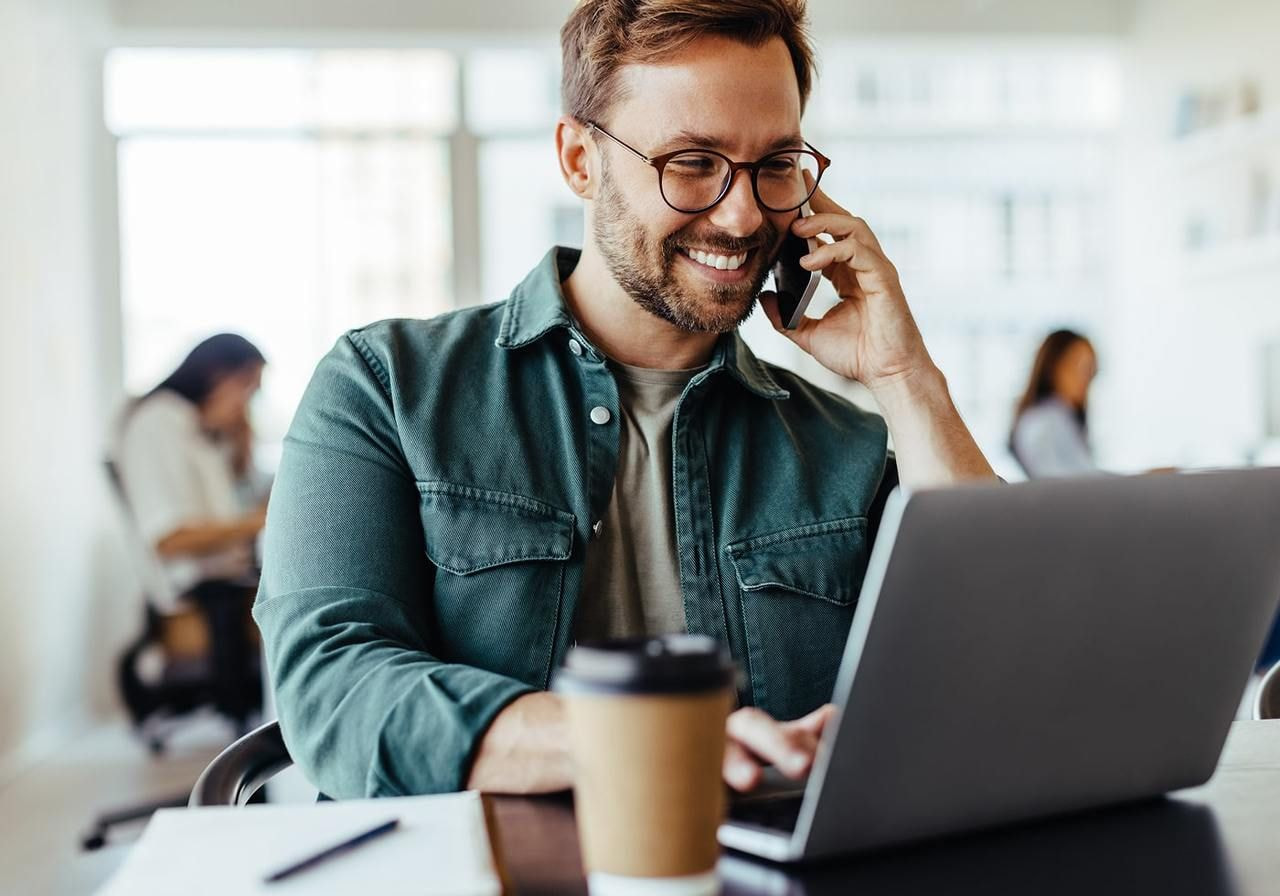A man with glasses smiles as he talks on the phone and works on a laptop in a bright office.