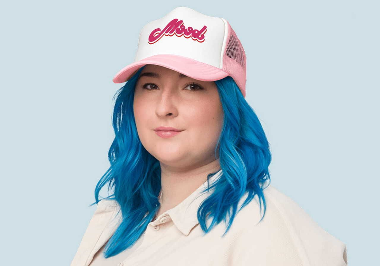 A woman with vibrant blue hair wearing a pink and white trucker hat displaying the word "Mood".