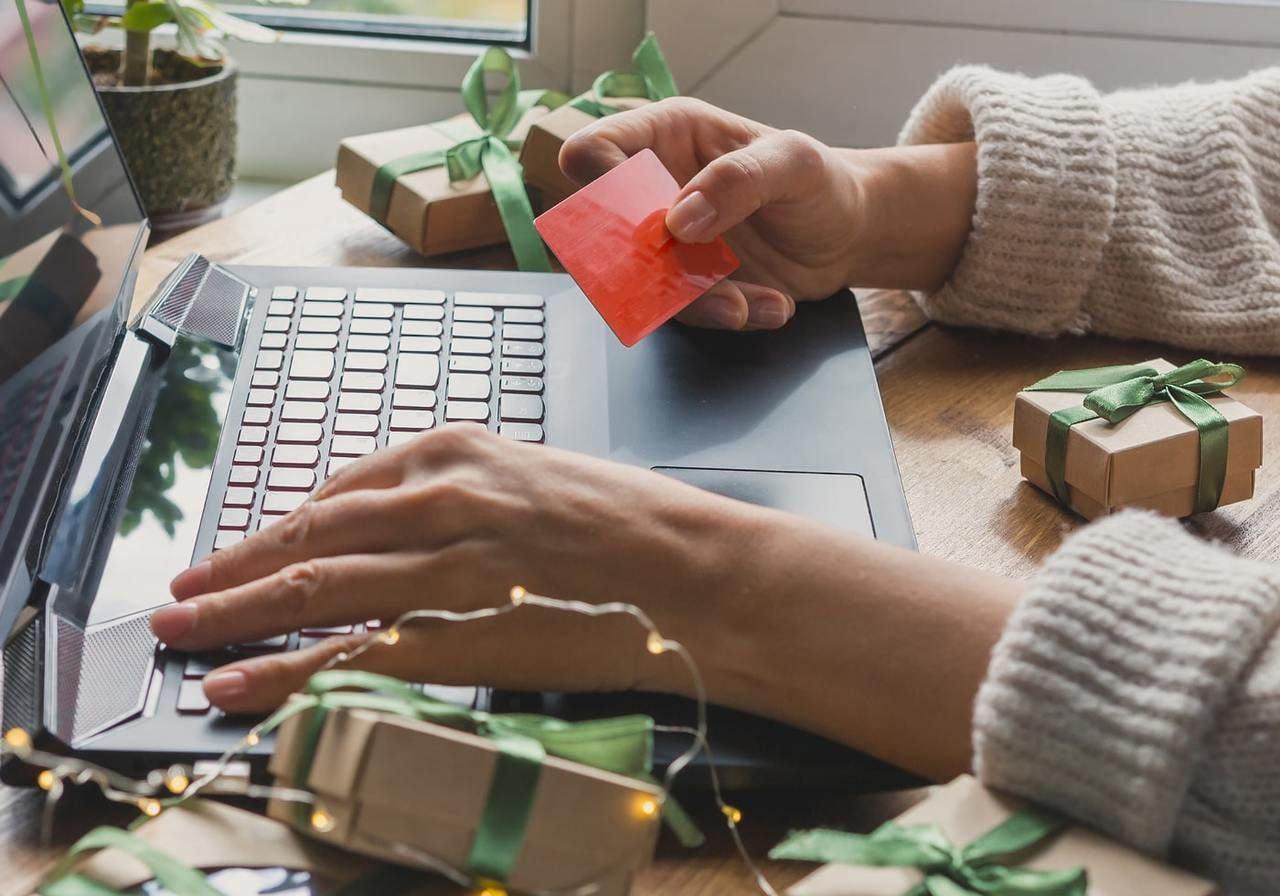A woman’s hands are holding a red credit card over a laptop, surrounded by small gift boxes with green ribbons and fairy lights.