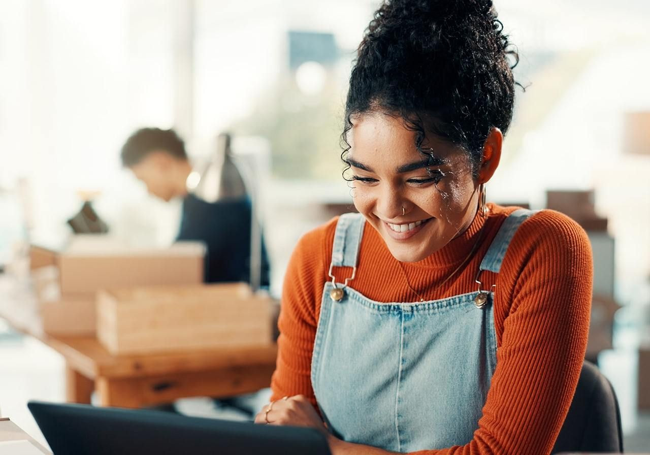 A woman in a denim pinafore and orange sweater smiles at a laptop in a bright office.