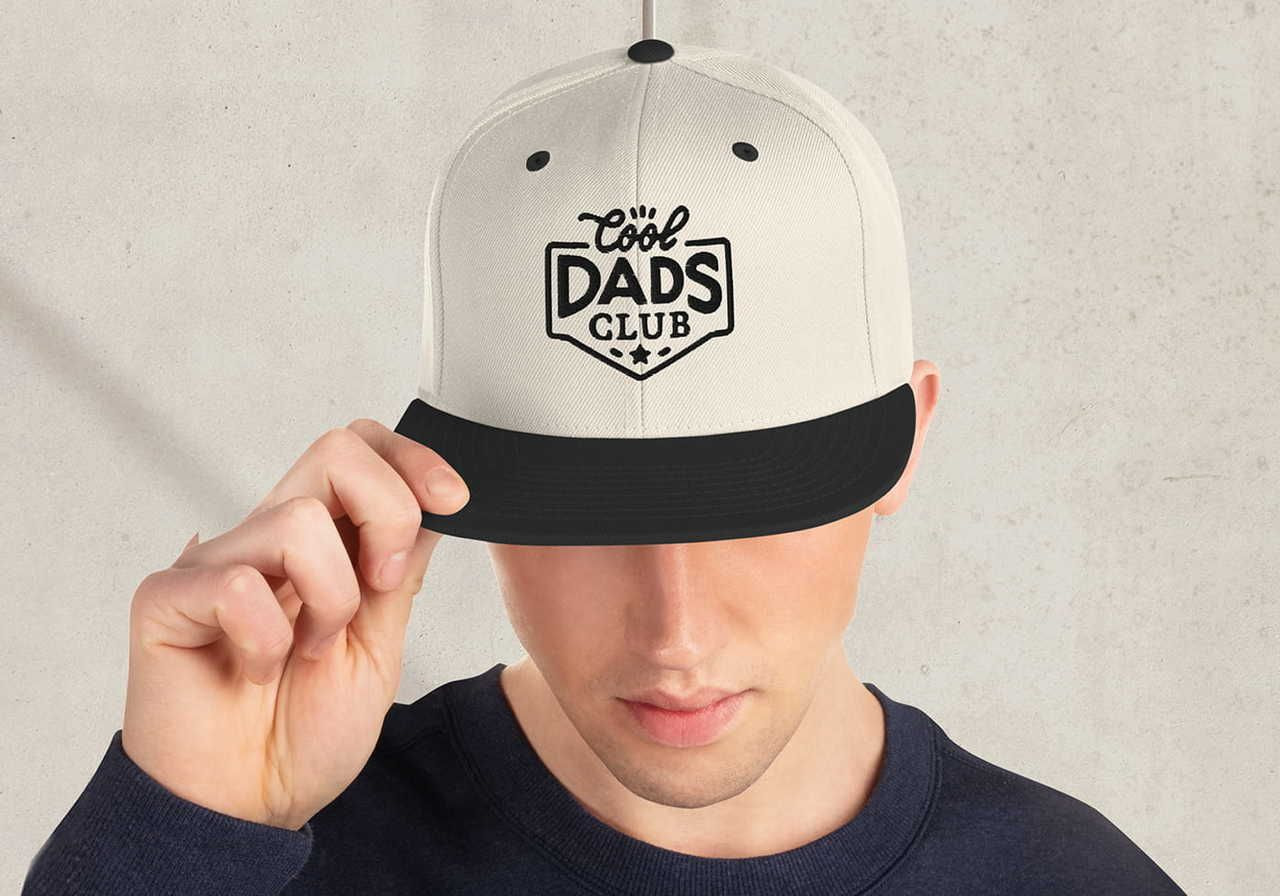 A man in a navy sweater adjusts a white baseball cap with a black brim, featuring the text "Cool Dads Club" in bold black letters.