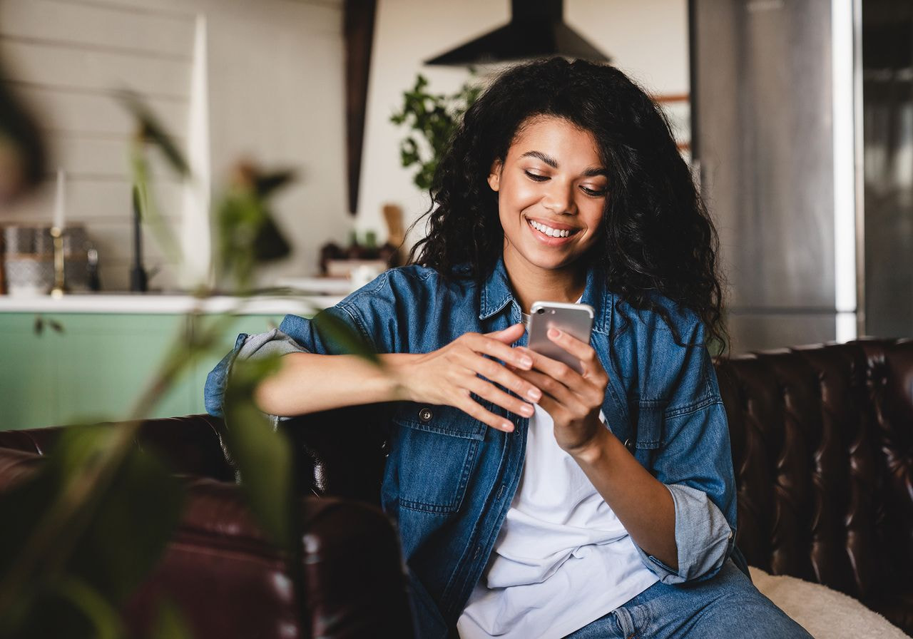 A woman in a denim shirt smiles while looking at her phone, seated on a leather sofa.