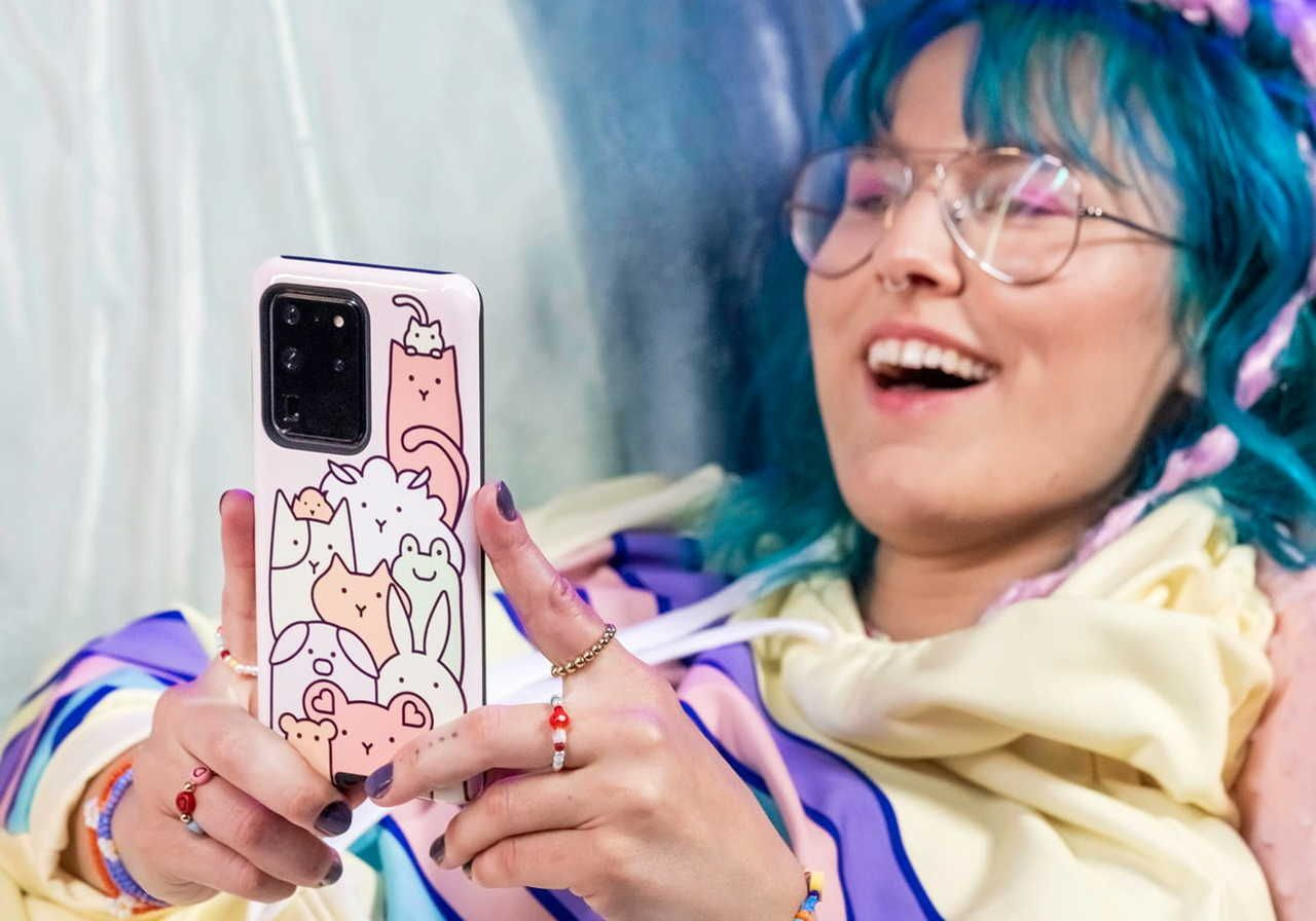 A young woman with blue hair and glasses holds a phone featuring a cute, colorful case while smiling.