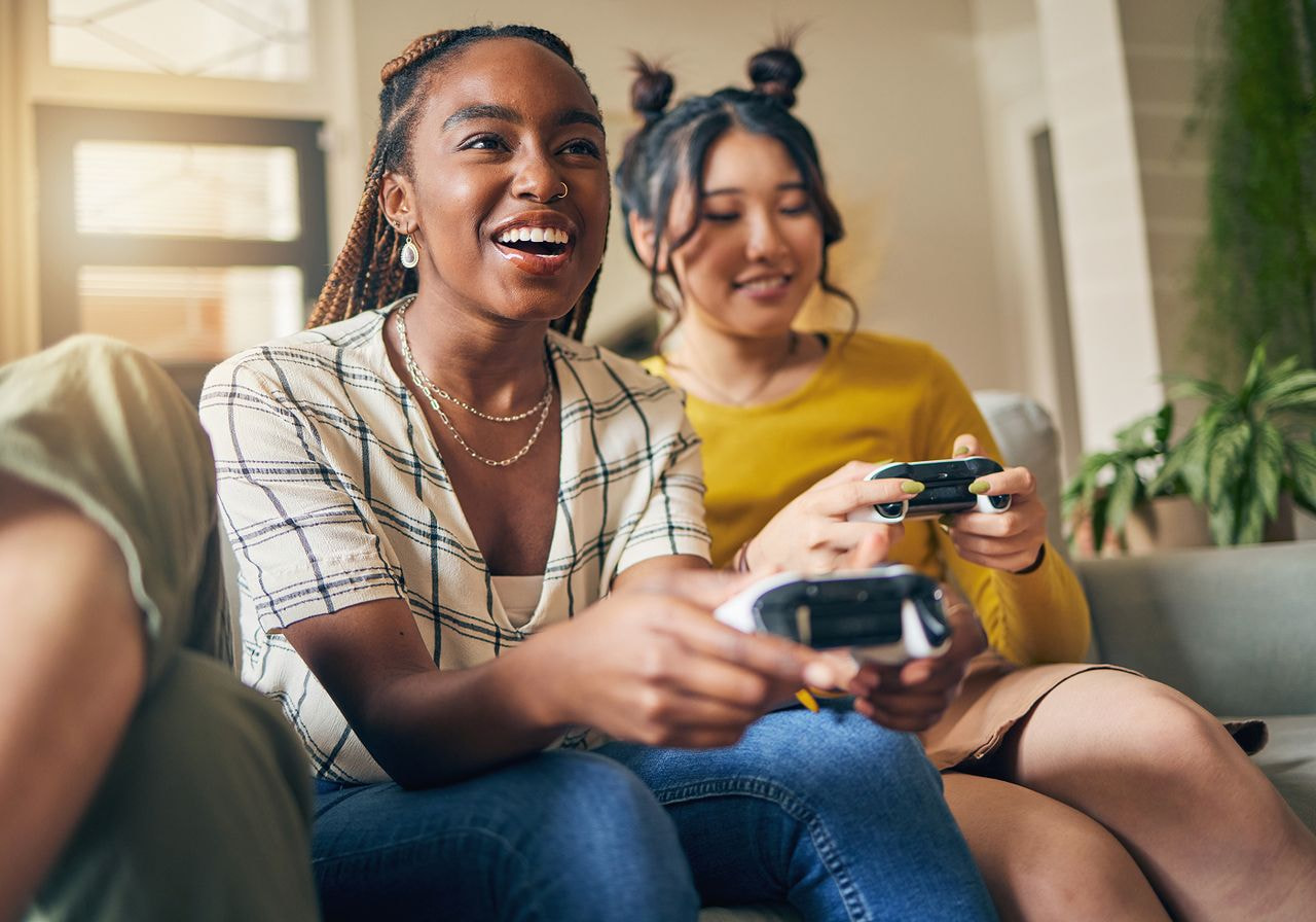 Two women sitting on a couch, focused on playing video games together, with controllers in hand and smiles on their faces.