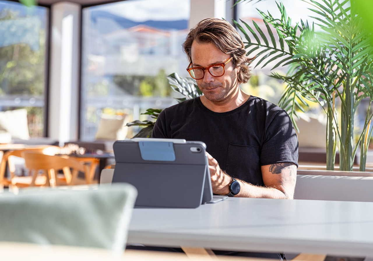 A man with glasses, wearing a black shirt, focused on a tablet, reading an article on “copyright vs trademark”, while in a cafe.