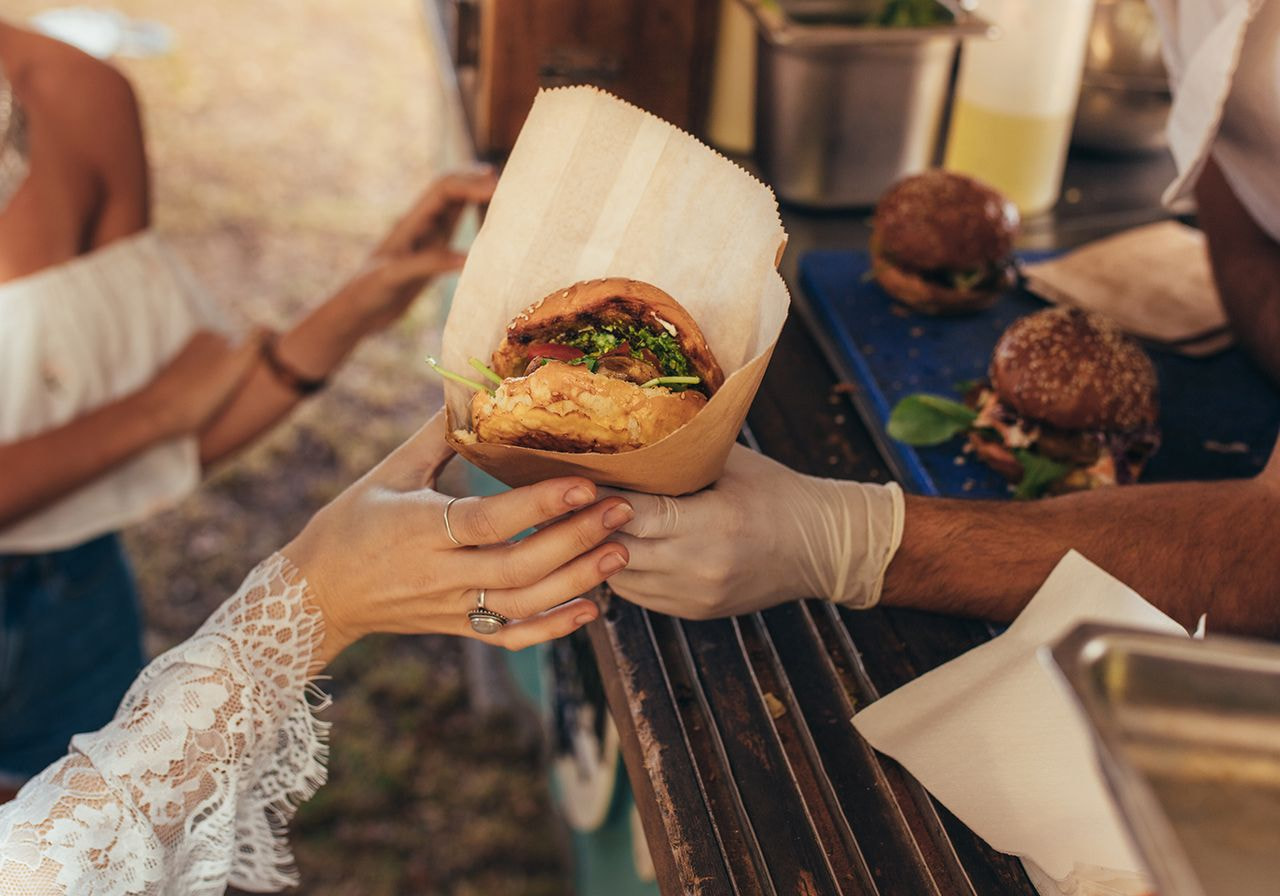 A person in lace sleeve hands a loaded burger wrapped in paper to a gloved food truck vendor.