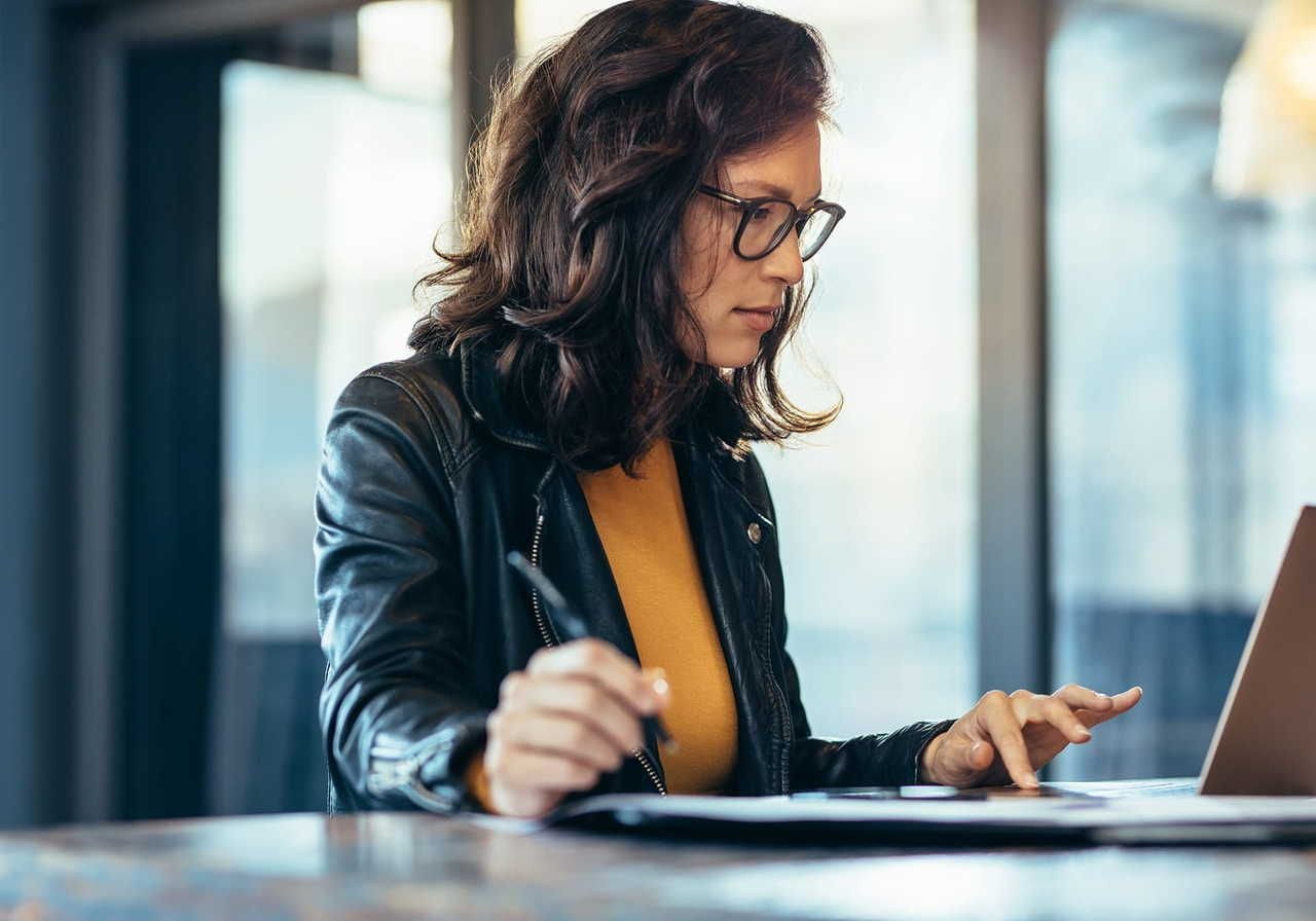 A woman wearing glasses, a yellow sweater, and a leather jacket is working on a laptop.