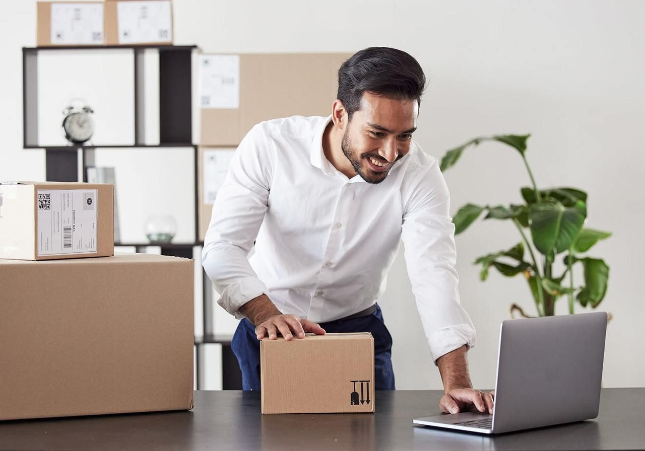 A smiling man is using a laptop with different delivery boxes in the background.