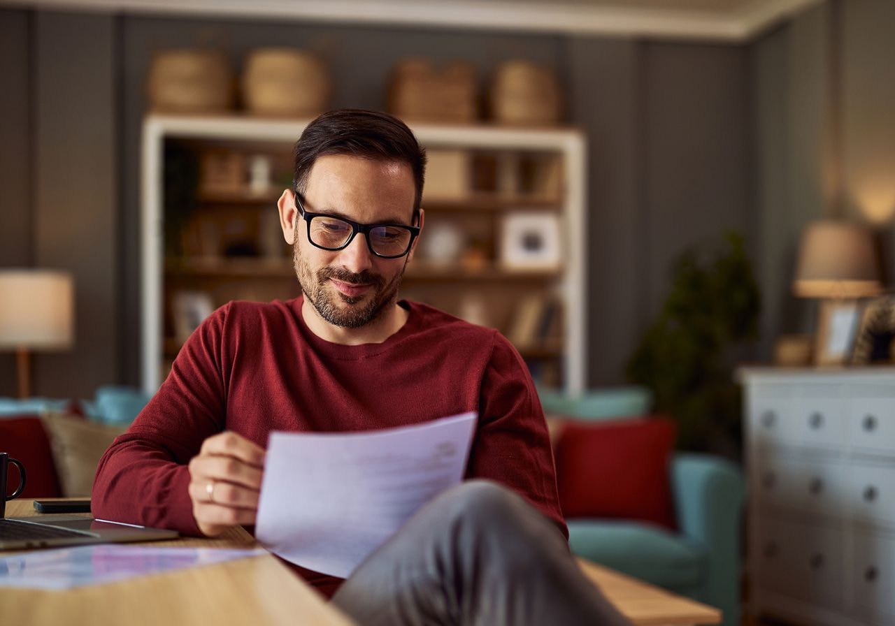 A man wearing glasses sits at a table, focused on a piece of paper in front of him.