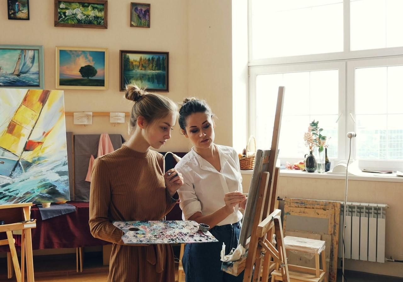 Two women stand beside an easel displaying a colorful painting, engaged in conversation about the artwork.