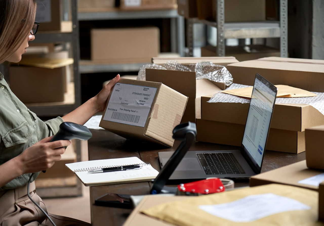 A woman in a warehouse scans a shipping label on a cardboard box. She sits at a table with a laptop, notepad, and packing materials nearby.