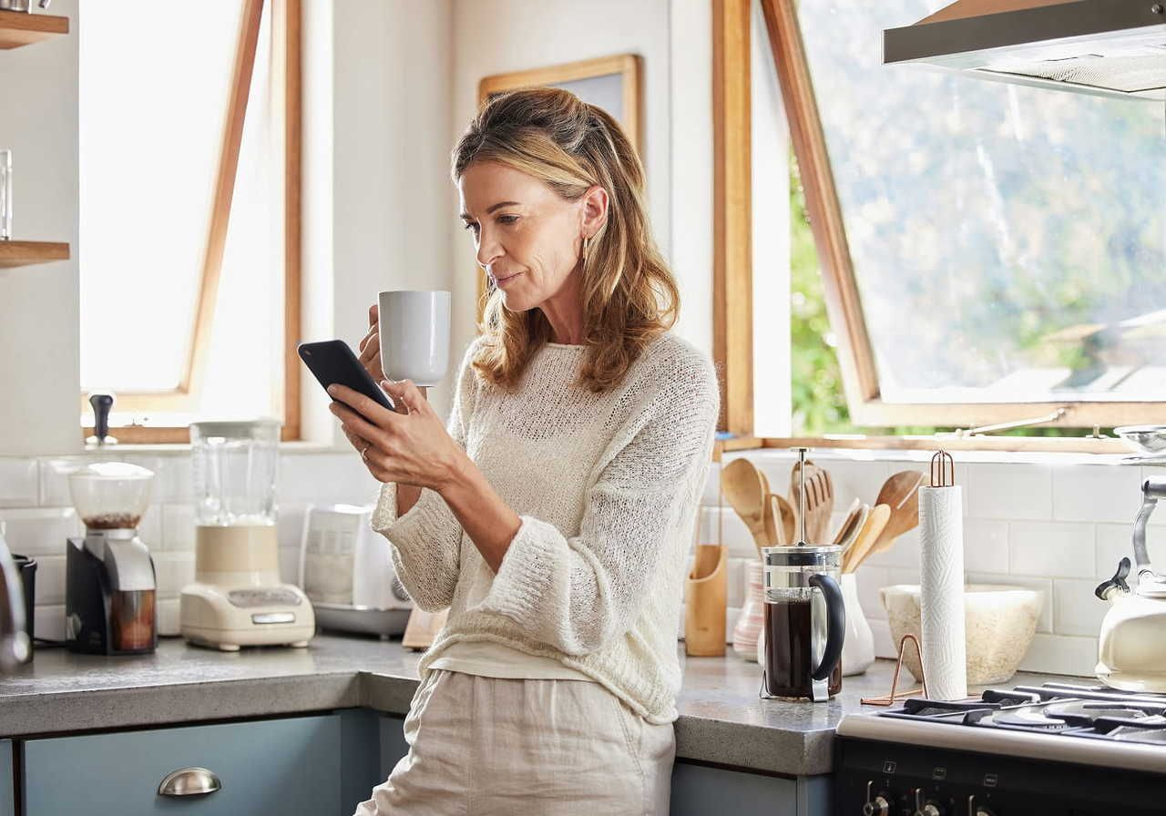 A woman in a light sweater is standing in a modern kitchen, looking at a phone in her hand and a coffee cup in the other.