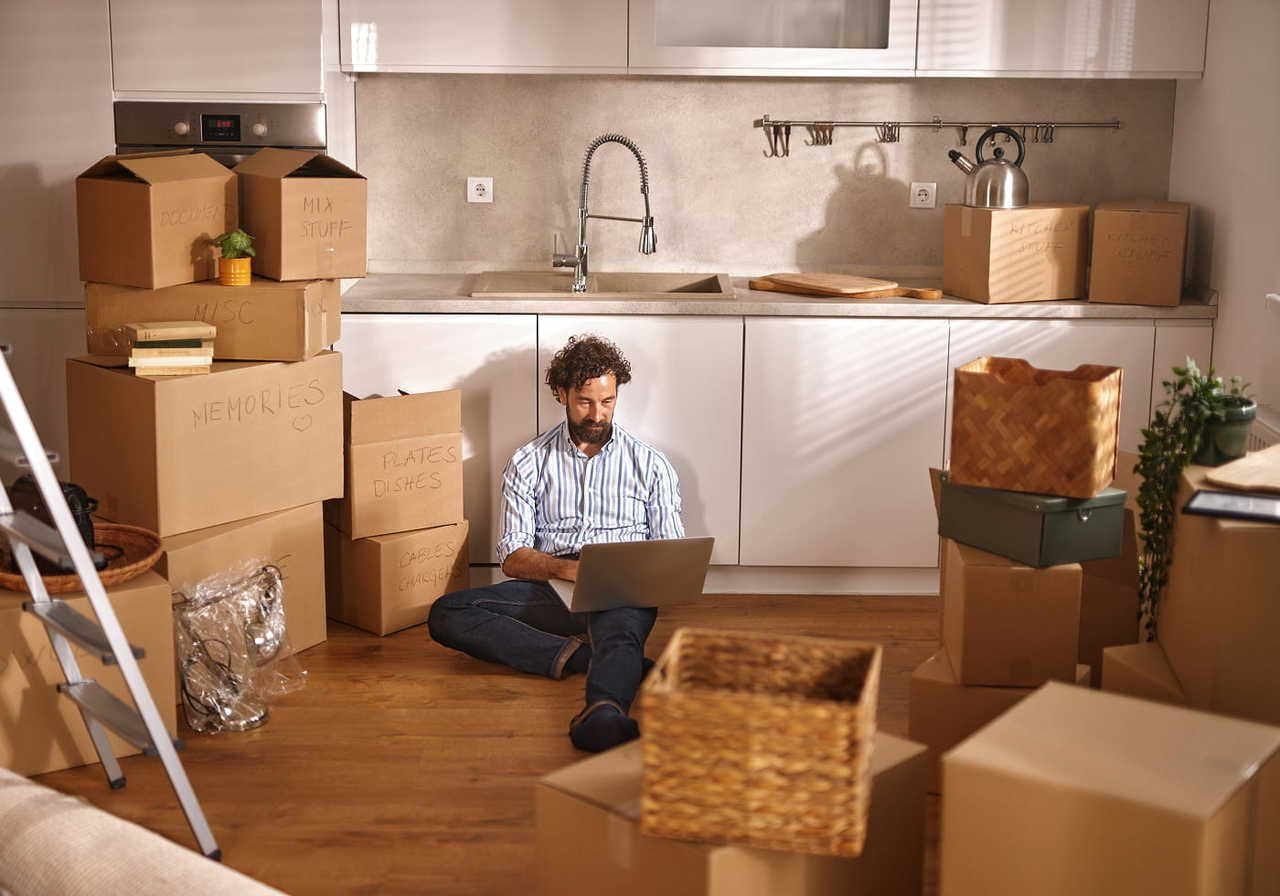 A man is sitting on a kitchen floor with a laptop surrounded by delivery boxes.